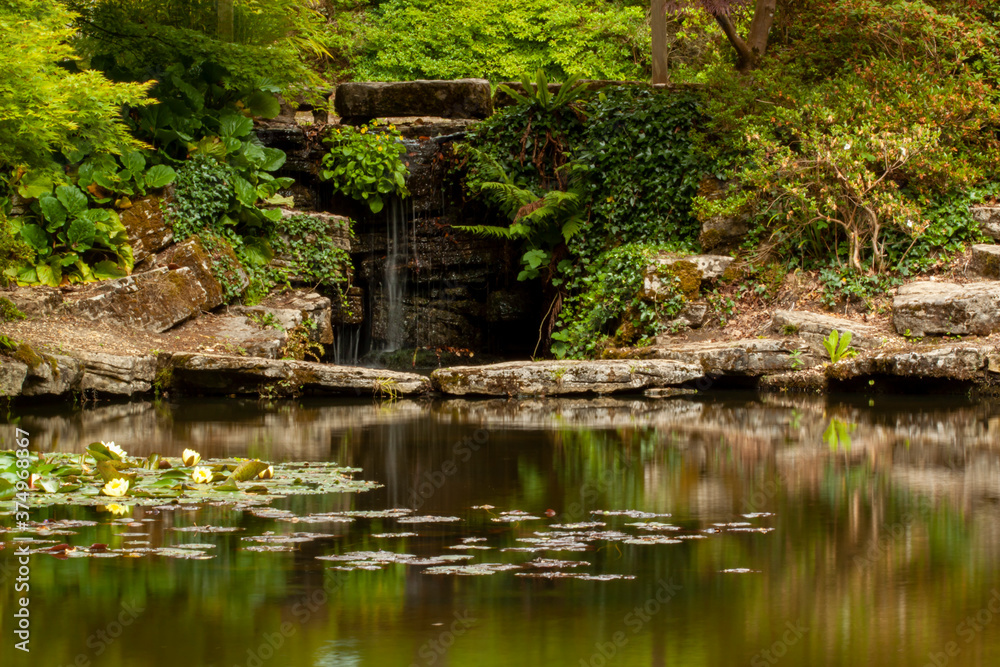 A long exposure image from a decorated backyard pond with water lilies and a small waterfall where water flows over smooth rocks into the pool. The area is shaded covered with trees, moss. A long exposure image from a decorated backyard pond with water lilies and a small waterfall where water flows over smooth rocks into the pool. The area is shaded covered with trees, moss.