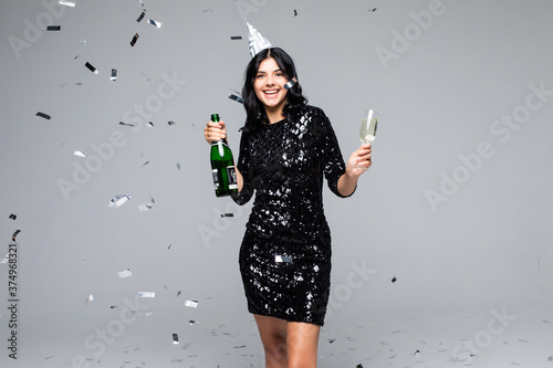 Portrait of an excited girl dressed in black dress holding bottle of champagne and a glass with a confetti rain isolated over gray background © F8  \ Suport Ukraine