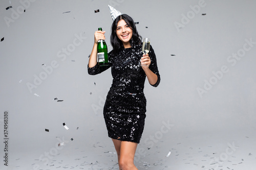 Young woman drinking champagne, celebrating new year, wearing black dress. Happy New Year to you. One young and beautiful woman dancing with glass of champagne and smiling on gray background © F8  \ Suport Ukraine