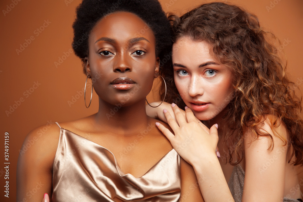 two pretty girls african and caucasian blond posing cheerful together on brown background, ethnicity diverse lifestyle people concept