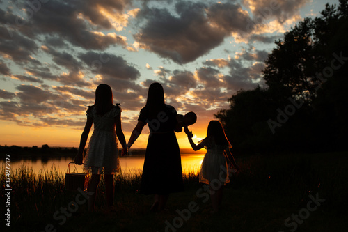 Silhouette of a happy family with children on nature. A mother is holding her seven-month-old son in her arms, daughters holding hands at sunset. They watch the pretty sunset with cloudy colourful sky © eduard