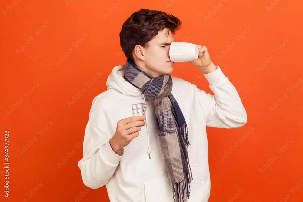 Sick upset teenager in casual style hoodie and checkered scarf taking medicine, suffering from cold or flu symptoms, healthcare. Indoor studio shot isolated on orange background Sick upset teenager in casual style hoodie and checkered scarf taking medicine, suffering from cold or flu symptoms, healthcare. Indoor studio shot isolated on orange background