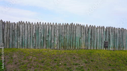 ancient fence made of wooden sticks, stockade, thick beams of pine tree with a sharp top, antique carpentry against a background of sky and green grass. palisade