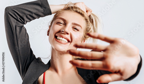 A beautiful blonde girl holds her hair and makes her own hairstyle with the emotion of happiness. Woman pulls her hand towards the camera on a gray isolated background for advertising. Nose piercing.