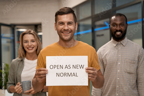 Office opening. Young happy man, office worker showing paper with text OPEN AS NEW NORMAL at camera and smiling while standing with colleagues in the office