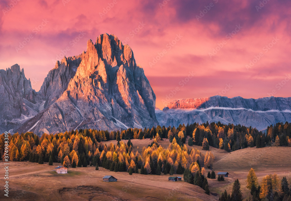 Beautiful mountains with lighted peaks and red sky at sunset. Autumn landscape with small wooden houses, mountain valley, meadows with green grass, fall trees, high rocks. Alpe di Siusi in Italy