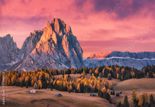 Beautiful mountains with lighted peaks and red sky at sunset. Autumn landscape with small wooden houses, mountain valley, meadows with green grass, fall trees, high rocks. Alpe di Siusi in Italy