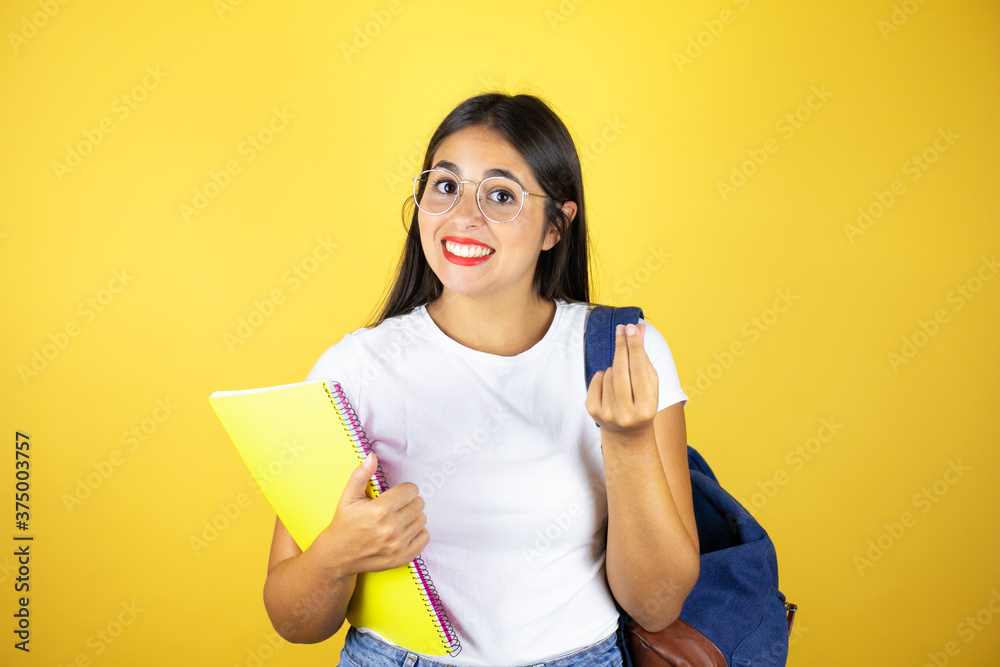 Young beautiful student woman wearing backpack holding notebook over isolated yellow background doing money gesture with hands, asking for salary payment, millionaire business