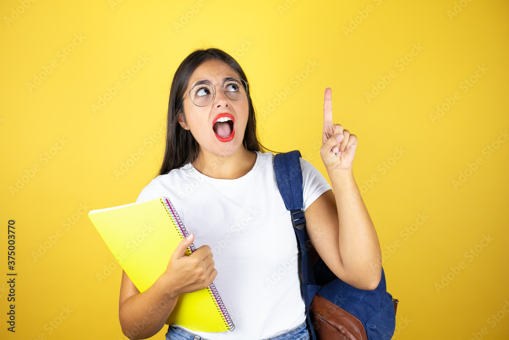 Young beautiful student woman wearing backpack holding notebook over isolated yellow background surprised, pointing and looking up. Young beautiful student woman wearing backpack holding notebook over isolated yellow background surprised, pointing and looking up.