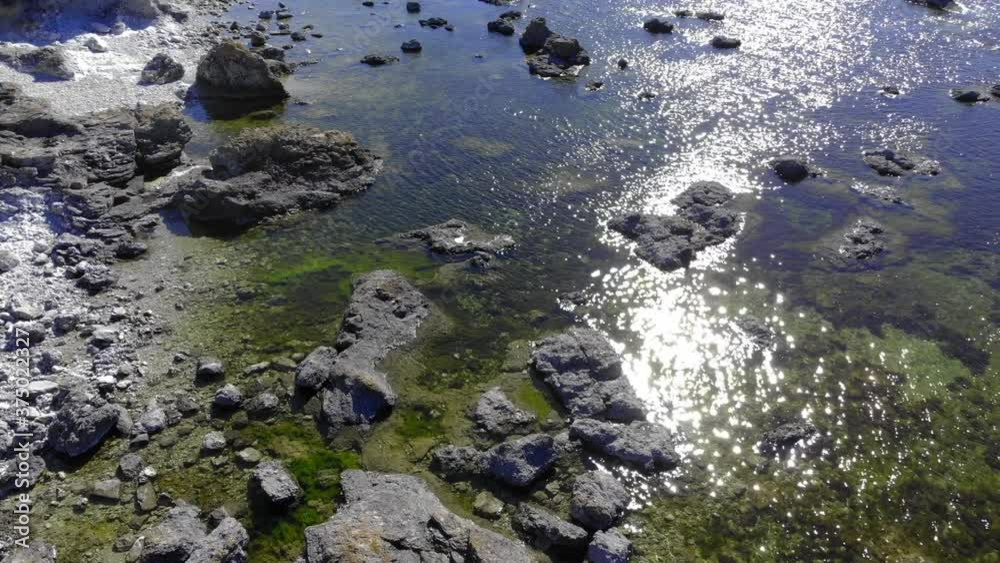 Aerial view over stones in shallow water, sun glittering on the water surface, on a warm, summer day, in Faro, Gotland, Sweden - low, tilt up,, drone shot