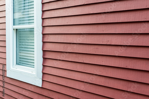 A small closed glass double hung window with white trim. There's a white blind hanging from the inside of the window. The exterior wall of the building is made of bright red narrow wooden clapboard. © Dolores  Harvey