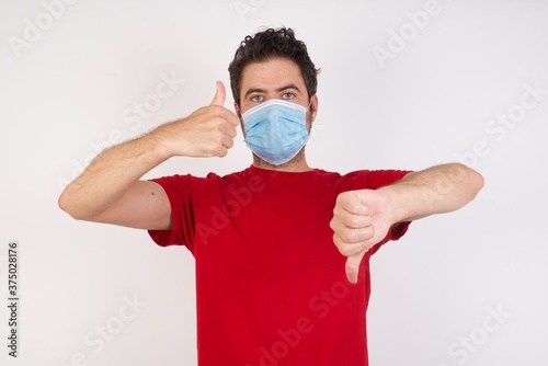 Young caucasian man with short hair wearing medical mask standing over isolated white background showing thumbs up and thumbs down, difficult choose concept