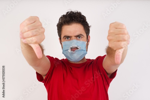 Young caucasian man with short hair wearing broken medical mask standing over isolated white background being upset showing thumb down with two hands. Dislike concept.