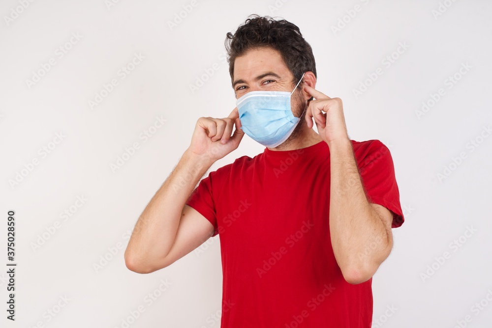 Young caucasian man with short hair wearing medical mask standing over isolated white background covering ears with fingers with annoyed expression for the noise of loud music. Deaf concept. Young caucasian man with short hair wearing medical mask standing over isolated white background covering ears with fingers with annoyed expression for the noise of loud music. Deaf concept.