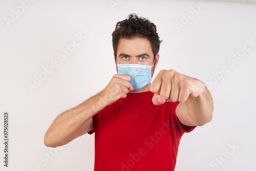 YoYoung caucasian man with short hair wearing medical mask standing over isolated white background Punching fist to fight, aggressive and angry attack, threat and violence