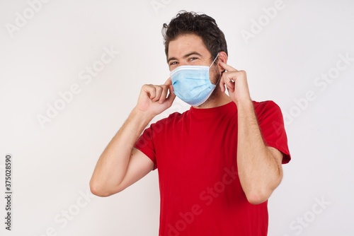 Young caucasian man with short hair wearing medical mask standing over isolated white background covering ears with fingers with annoyed expression for the noise of loud music. Deaf concept.