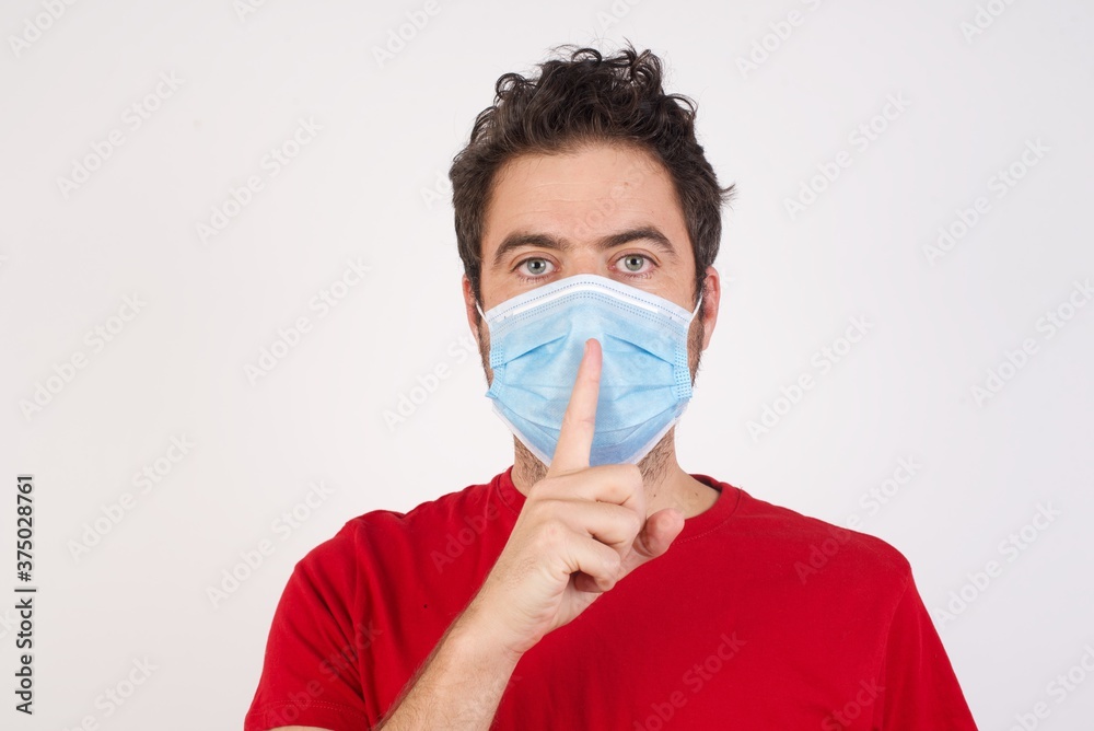 Young caucasian man with short hair wearing medical mask standing over isolated white background asking to be quiet with finger on lips. Silence and secret concept.