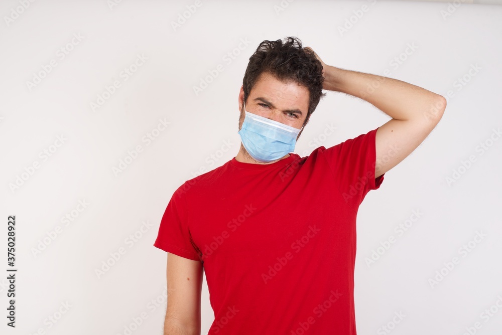 Young caucasian man with short hair wearing medical mask standing over isolated white wall being confused and wonders about something. Holding hand on her head, uncertain with doubt. Pensive concept. Young caucasian man with short hair wearing medical mask standing over isolated white wall being confused and wonders about something. Holding hand on her head, uncertain with doubt. Pensive concept.