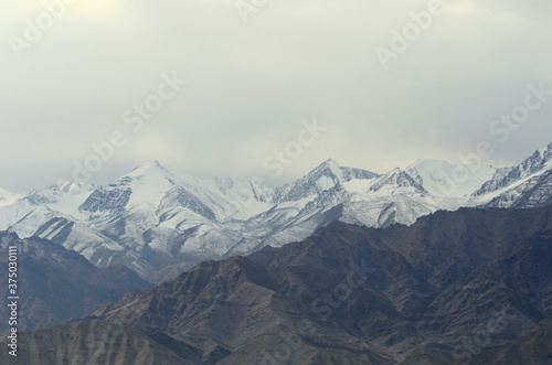 A mountain range, part of the Western Himalayas in Ladakh, India, is covered in snow. The sky is filled with snow clouds. A lower rocky mountain slope is in the foreground.