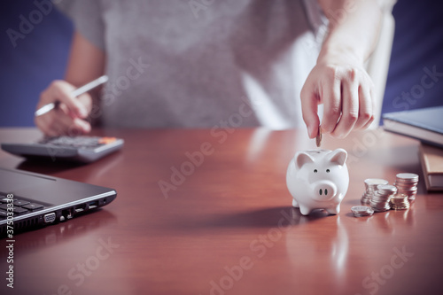 Female hand putting money into piggy bank with using calculator to calculate and money stack, for saving money wealth and financial, business, investment, retirement, saving for the future concept.