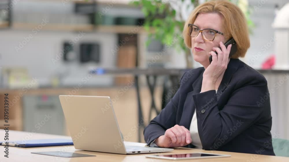 Old Businesswoman with Laptop Talking on Smartphone in Office Old Businesswoman with Laptop Talking on Smartphone in Office
