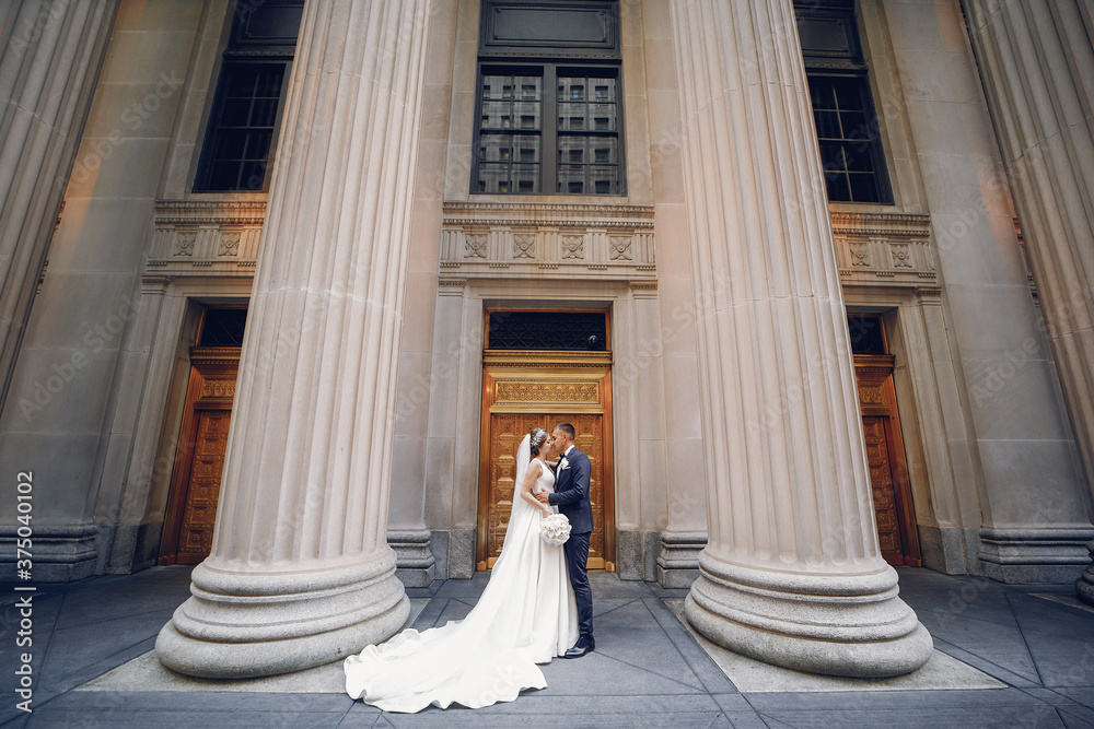 Elegant bride in a white dress and veil. Handsome groom in a blue suit. Couple near large building with columns