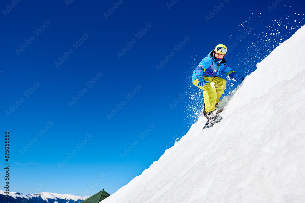 Male snowboarder riding snowboard fast down steep snowy mountain slope, jumping in air on copy space background of blue sky and white snow on sunny winter day. Extreme sport and recreation concept.