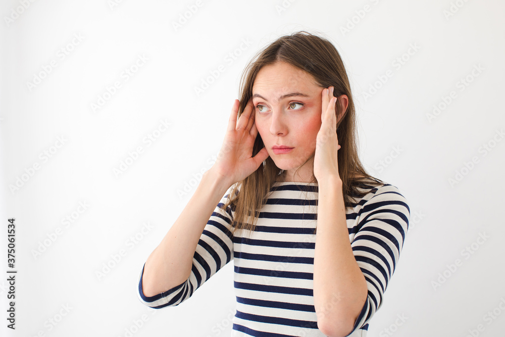 Young female in casual striped wear having headache and rubbing temples while standing against white background