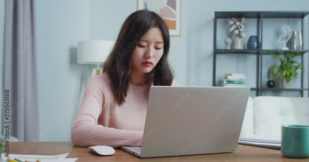 Charming Asian woman sitting at table and working in home office room. Concentrated female worker typing, shutting laptop computer and looking at camera. Self-isolation, occupation, job. Charming Asian woman sitting at table and working in home office room. Concentrated female worker typing, shutting laptop computer and looking at camera. Self-isolation, occupation, job.