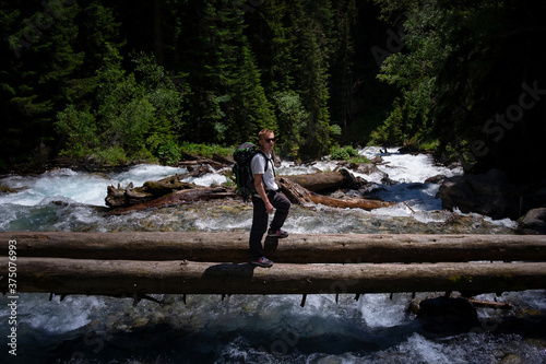 Hiker man hiking crossing river walking in balance on fallen tree trunk