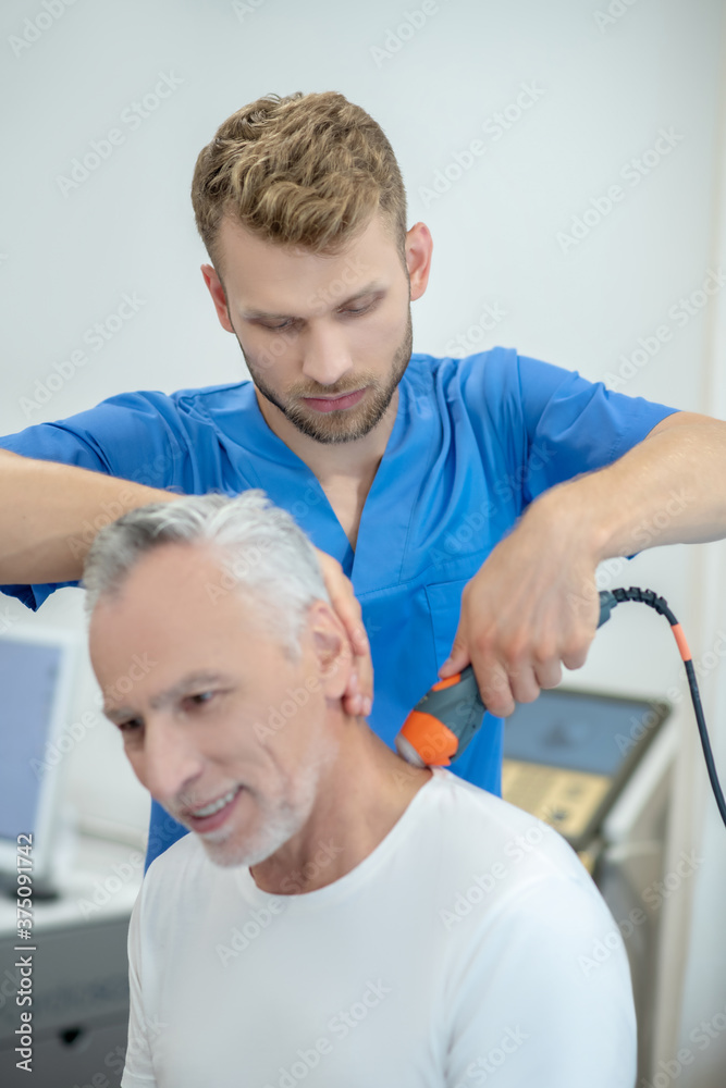 Fototapeta premium Bearded doctor performing ultrasound neck treatment on smiling mature patient