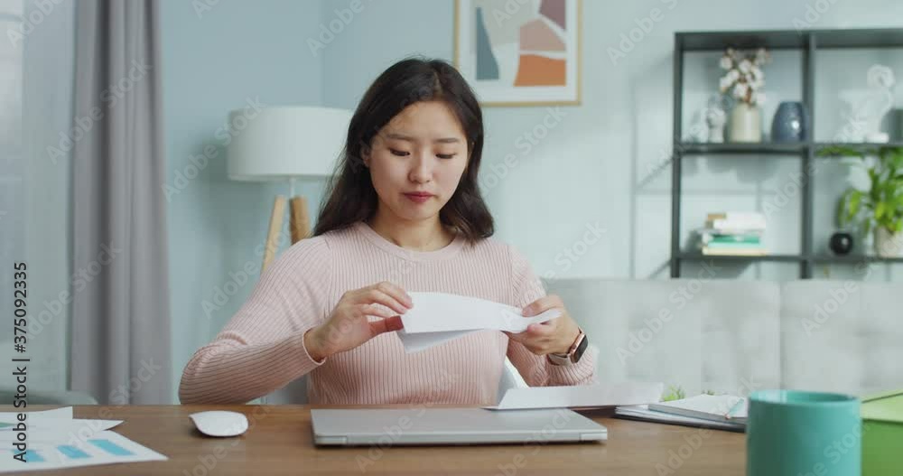 Good-looking Asian girl siitting at table and opening letter from university, college at home. Emotional young woman reading respose and being sad, disappointed. Education, admission, profession. Good-looking Asian girl siitting at table and opening letter from university, college at home. Emotional young woman reading respose and being sad, disappointed. Education, admission, profession.