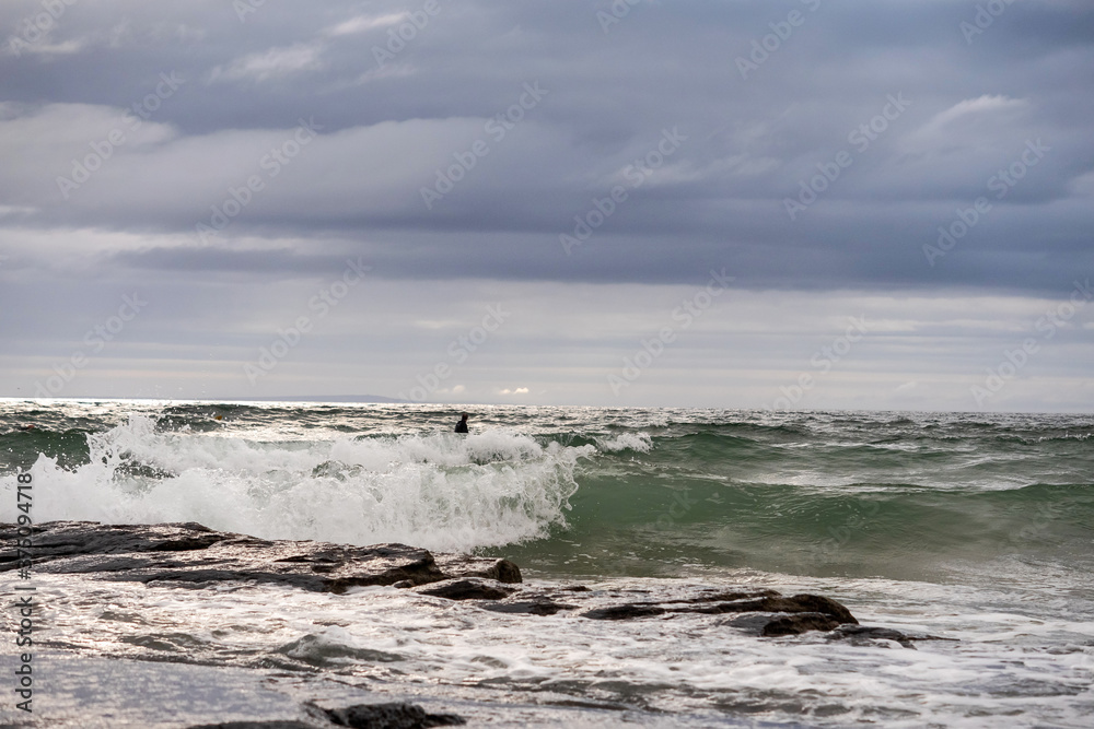 Powerful ocean waves and surfer in black wet suit, Cloudy sky.