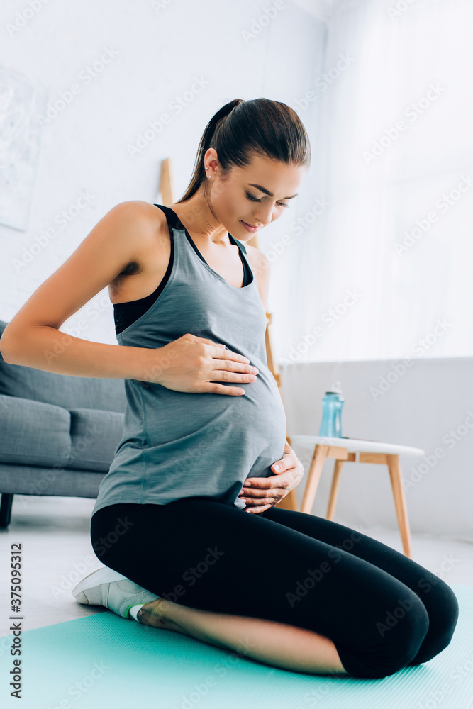 Pregnant sportswoman touching belly while sitting on fitness mat in living room Pregnant sportswoman touching belly while sitting on fitness mat in living room