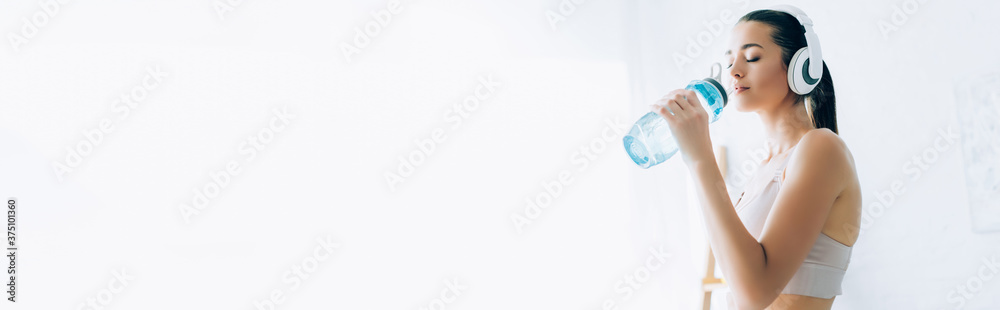 Panoramic shot of sportswoman in headphones drinking water at home Panoramic shot of sportswoman in headphones drinking water at home