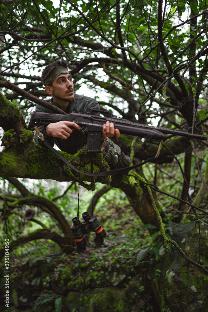 Soldier or revolutionary member or hunter in camouflage on the tree observing the gun in his hand Soldier or revolutionary member or hunter in camouflage on the tree observing the gun in his hand