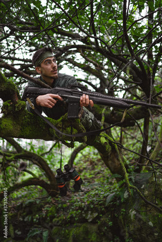 Soldier or revolutionary member or hunter in camouflage on the tree observing the gun in his hand