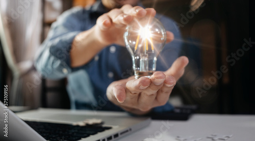 business hand holding lightbulb with using smartphone, laptop computer and money stack in office. idea saving energy and accounting finance concept in morning light