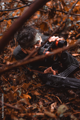 Soldier or revolutionary member or hunter aiming with gun and binocular in his hands in camouflage in the forest floor, hunt concept