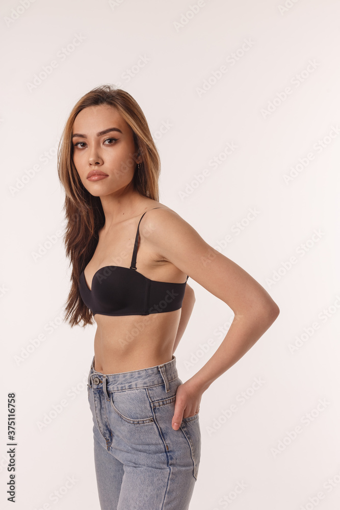 Foto de portrait of sexy asian woman with long hair, stylish earrings  posing in black lingerie and blue jeans isolated on white background. model  tests of skinny girl in bra. attractive female
