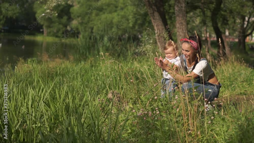 Mother with baby son sitting near pond in park. Toddler trying to clap hands together with mom, looking around. Child experiencing beauty of nature: green trees, birds and water. Concept of motherhood