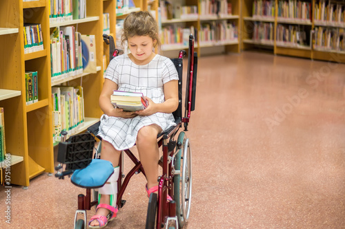 Caucasian little girl sitting in wheelchair at the library. The disabled teenager is choosing books