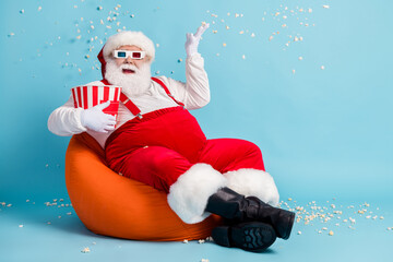 Full length photo of pensioner old man watch blockbuster throw popcorn make mess sit orange beanbag wear red santa x-mas costume suspender 3d glasses cap boot isolated blue color background
