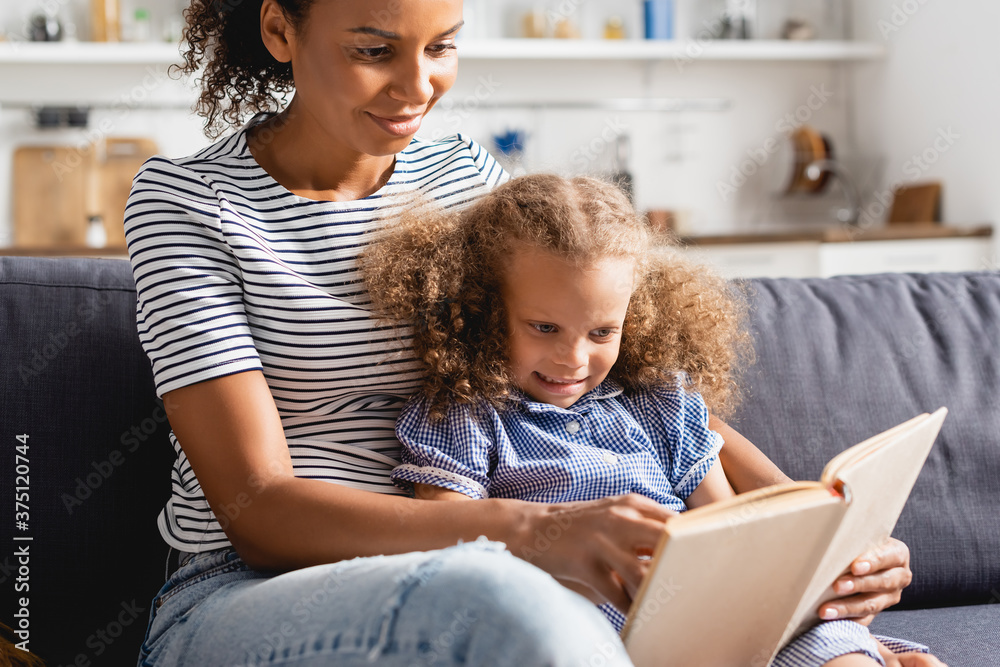 selective focus of african american nanny and girl sitting on sofa and reading book together selective focus of african american nanny and girl sitting on sofa and reading book together