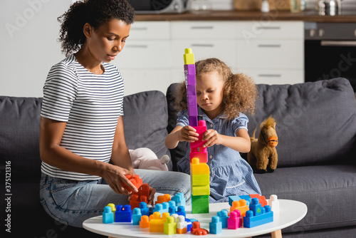 african american kid building tower from multicolored blocks while sitting on couch near nanny