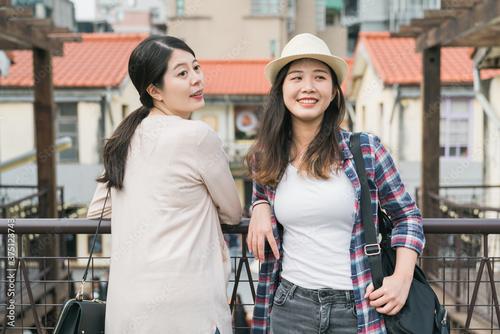 Two smiling young women friends dressed casually spending time together travel in old town. girl tourists leaning on rail while standing and talking look at same way outdoors. real moments lifestyle