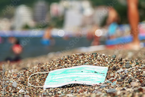 Discarded used face mask lies on a pebble beach, beachgoers are relaxing by the sea in the background. Becici, Budva Municipality, Montenegro