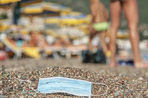 Discarded used facial mask lies on a sandy pebble beach, in the background beachgoers are relaxing by sun loungers and beach parasols. Becici, Budva Municipality, Montenegro