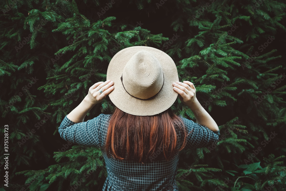 Portrait stylish red-haired young woman in a straw hat and dress on a background of green trees. Shoot from the back. Place for text or advertising