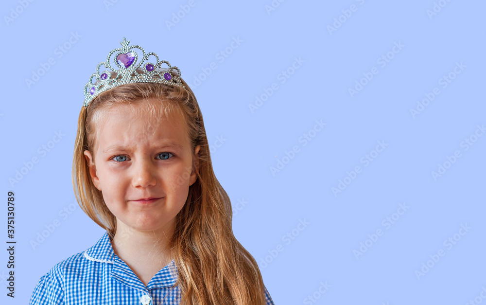 Beautiful little princess girl in silver crown looking at the camera and crying. Serious emotional young lady with long wavy hair in plaid dress portrait on pastel blue background with copy space. Beautiful little princess girl in silver crown looking at the camera and crying. Serious emotional young lady with long wavy hair in plaid dress portrait on pastel blue background with copy space.